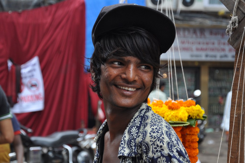 Close-up of a smiling young man with long hair, wearing a black cap and a patterned shirt, standing in a bustling market area with colorful flower garlands in the background.